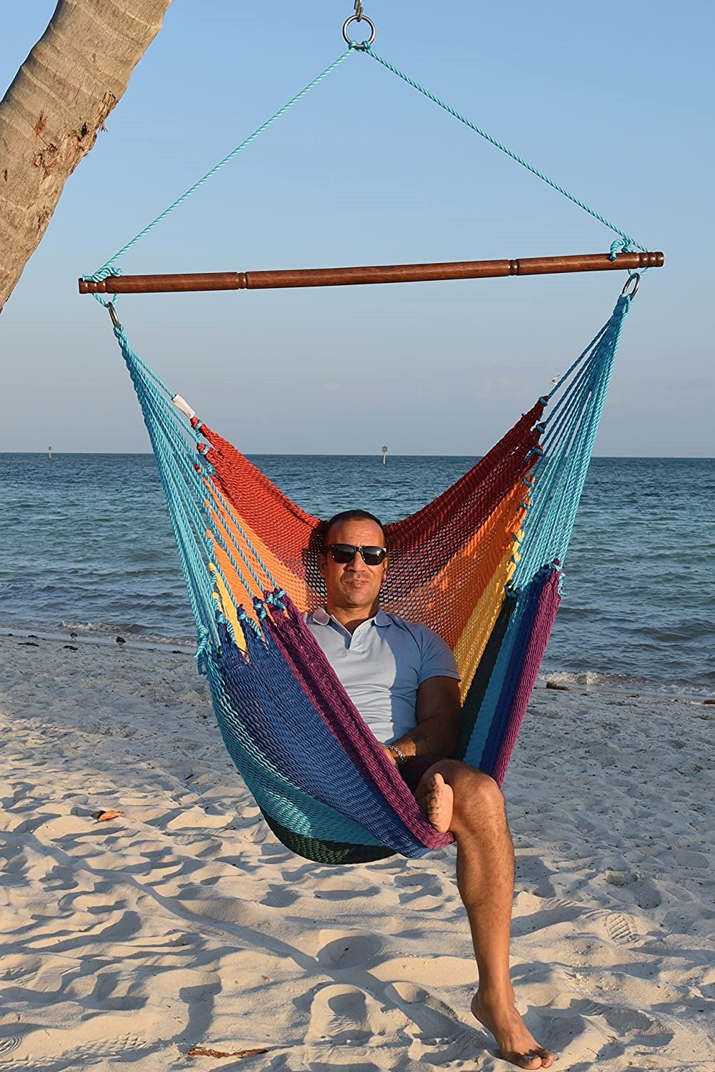 Man in sunglasses seated in a rainbow Jumbo Caribbean Hammock Chair on a white sand beach