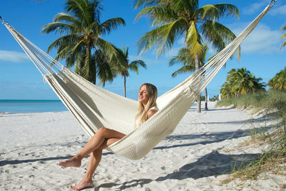 Cream Jumbo Mayan Hammock strung between palm trees on a white-sand beach, a woman sitting in it with her feet on the sand