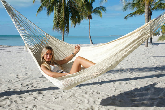 Woman reclining in the Cream Jumbo Mayan Hammock on a palm-lined beach, the full hand-woven bed in view