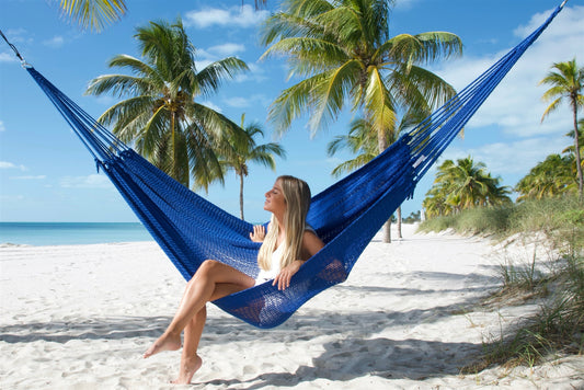 Dark Blue Jumbo Mayan Hammock strung between palm trees on a white-sand beach, a woman sitting in it with her feet on the sand