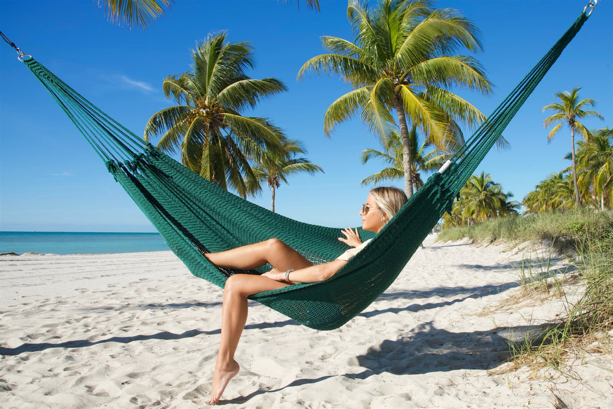 Green Jumbo Mayan Hammock strung between palm trees on a white-sand beach, a woman sitting in it with one foot on the sand