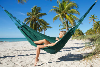 Green Jumbo Mayan Hammock strung between palm trees on a white-sand beach, a woman sitting in it with one foot on the sand
