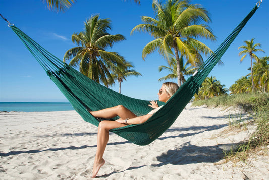 Green Jumbo Mayan Hammock strung between palm trees on a white-sand beach, a woman sitting in it with one foot on the sand
