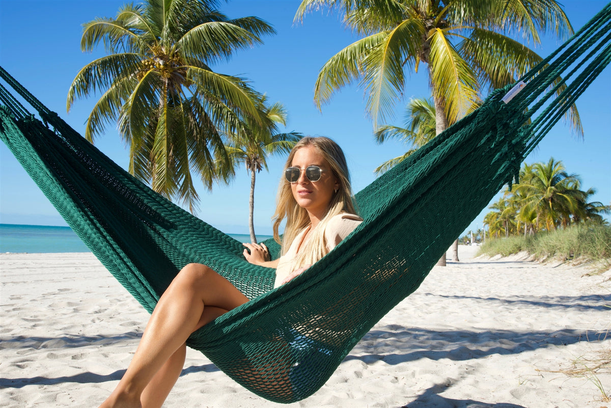 Woman sitting upright in the Green Jumbo Mayan Hammock on the beach, palm trees behind