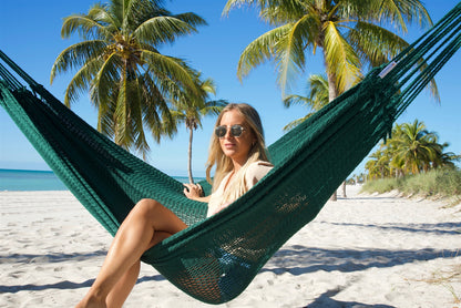 Woman sitting upright in the Green Jumbo Mayan Hammock on the beach, palm trees behind