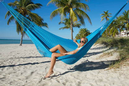 Woman sitting sideways in the Light Blue Jumbo Mayan Hammock with one foot on the sand, palm trees behind