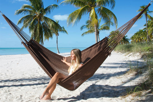 Mocha Jumbo Mayan Hammock strung between palm trees on a white-sand beach, a woman sitting in it with her feet on the sand