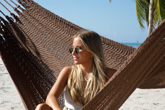 Close view of a woman in sunglasses sitting in the Mocha Jumbo Mayan Hammock, the hand-woven polyester bed framing her