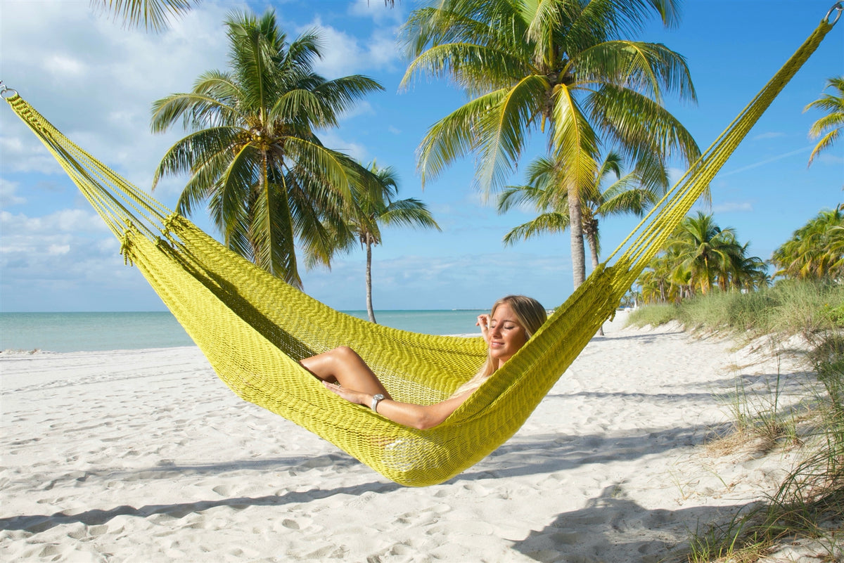 Olive Jumbo Mayan Hammock strung between palm trees on a white-sand beach, a woman reclining in the hand-woven polyester bed