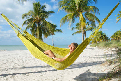 Olive Jumbo Mayan Hammock strung between palm trees on a white-sand beach, a woman reclining in the hand-woven polyester bed