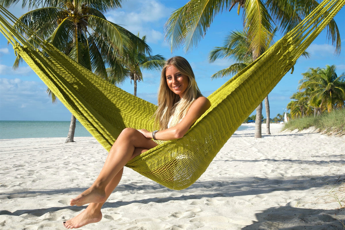 Woman sitting sideways in the Olive Jumbo Mayan Hammock with her feet toward the sand, palm trees behind