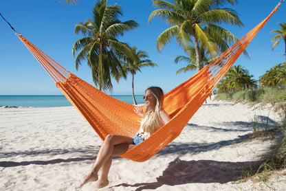 Orange Jumbo Mayan Hammock hung between palm trees on the beach, a woman sitting in it and glancing to the side