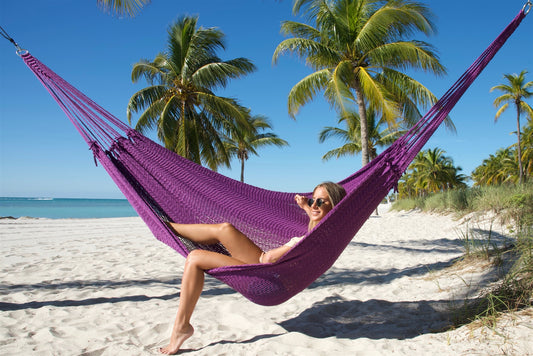 Purple Jumbo Mayan Hammock strung between palm trees on a white-sand beach, a woman sitting in it with one foot on the sand