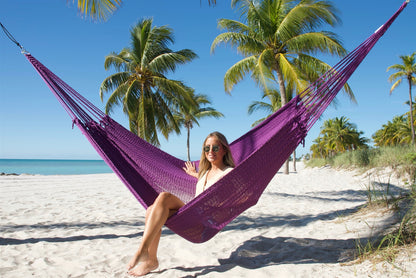 Woman sitting sideways in the Purple Jumbo Mayan Hammock with one foot on the sand, palm trees behind