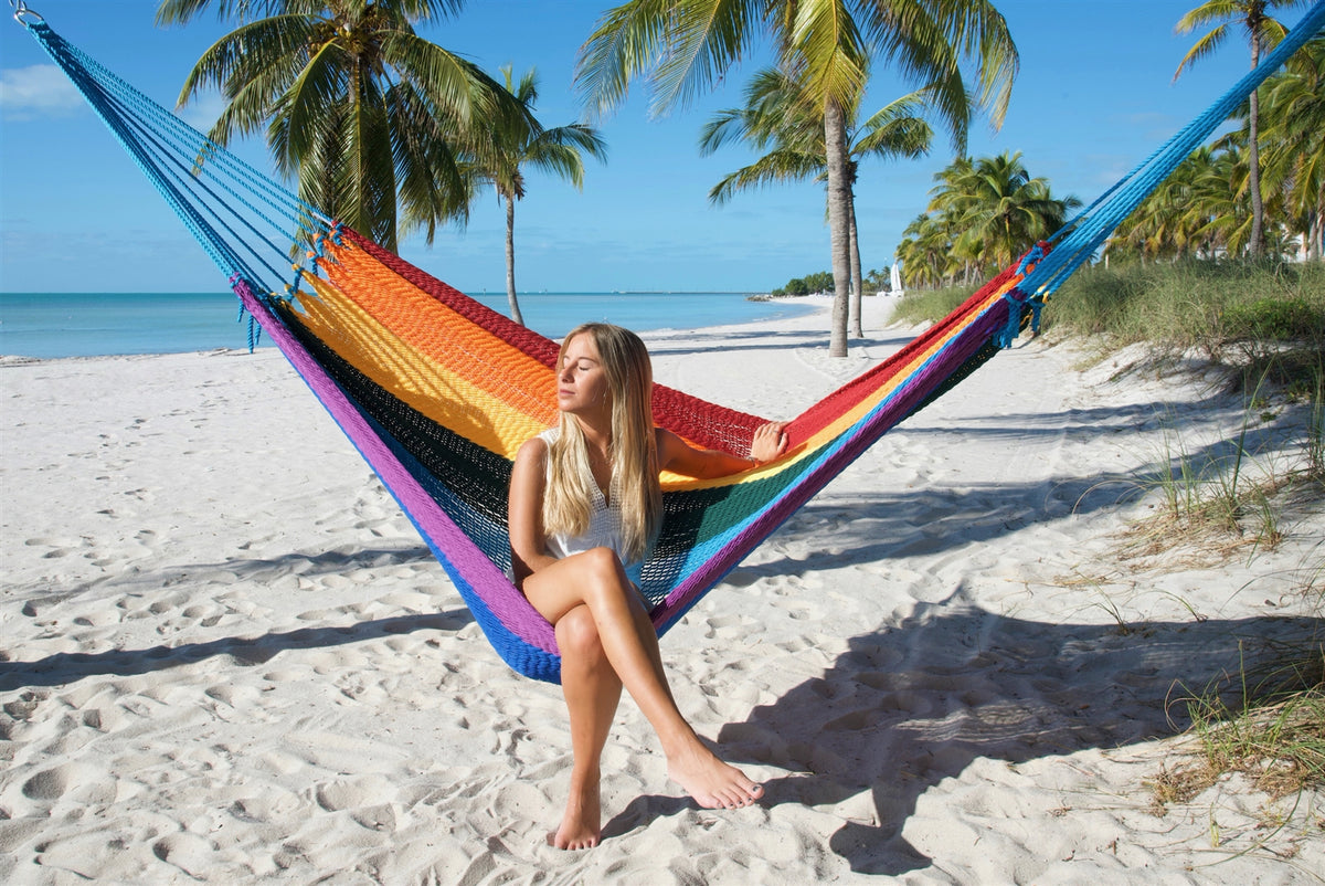 Woman sitting upright in the Rainbow Jumbo Mayan Hammock with her feet on the sand, palm trees behind