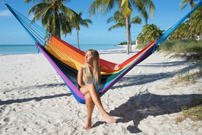 Woman sitting upright in the Rainbow Jumbo Mayan Hammock with her feet on the sand, palm trees behind