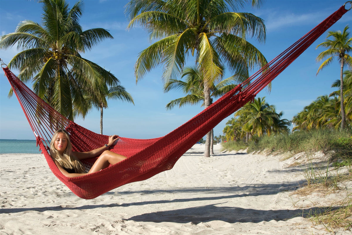 Red Jumbo Mayan Hammock strung between palm trees on a white-sand beach, a woman reclining in the hand-woven polyester bed