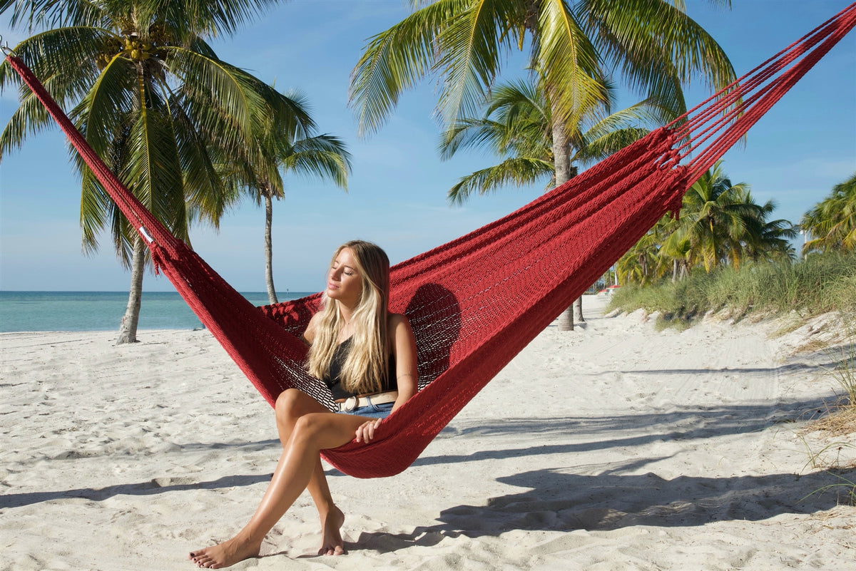 Woman sitting sideways in the Red Jumbo Mayan Hammock with her feet on the sand, palm trees behind