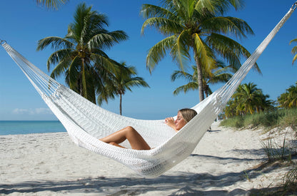 White Jumbo Mayan Hammock strung between palm trees on a white-sand beach, a woman reclining in the hand-woven polyester bed