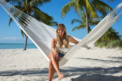 Woman sitting in the White Jumbo Mayan Hammock with one foot on the sand, palm trees behind