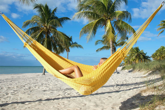 Yellow Jumbo Mayan Hammock strung between palm trees on a white-sand beach, a woman reclining in the hand-woven polyester bed