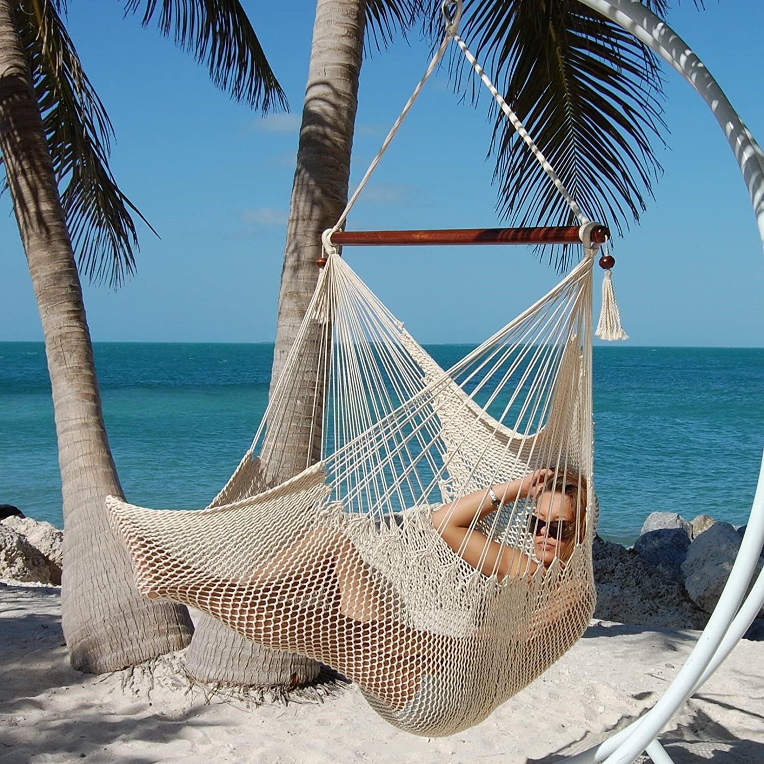 Woman reclining in a cream Large Caribbean Hammock Chair on a white arc stand beside palm trees and the ocean