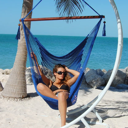 Woman seated in a dark blue Large Caribbean Hammock Chair on a white arc stand beside palm trees and the ocean