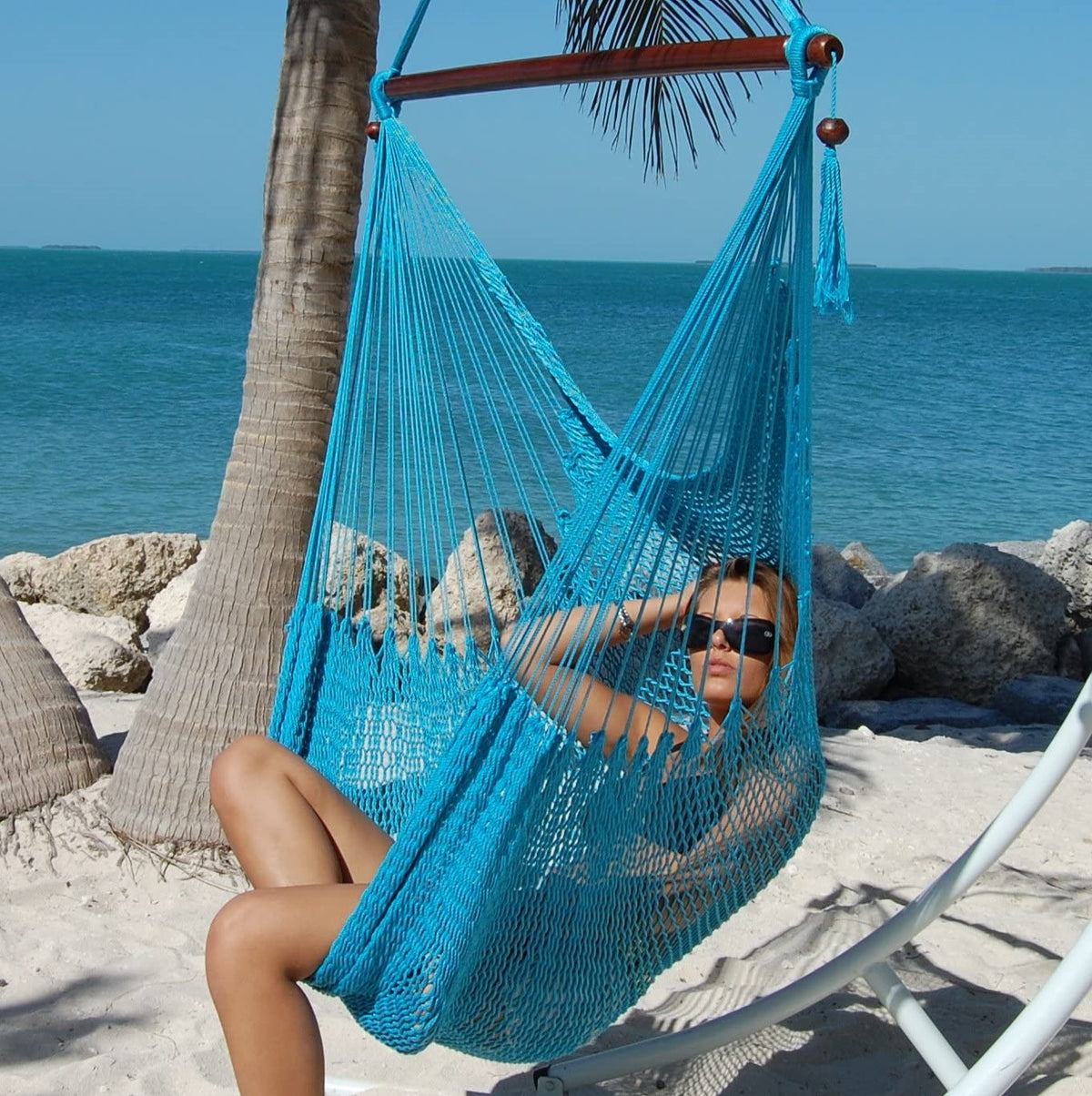 Woman reclining in a light blue Large Caribbean Hammock Chair on a white arc stand beside palm trees and the ocean