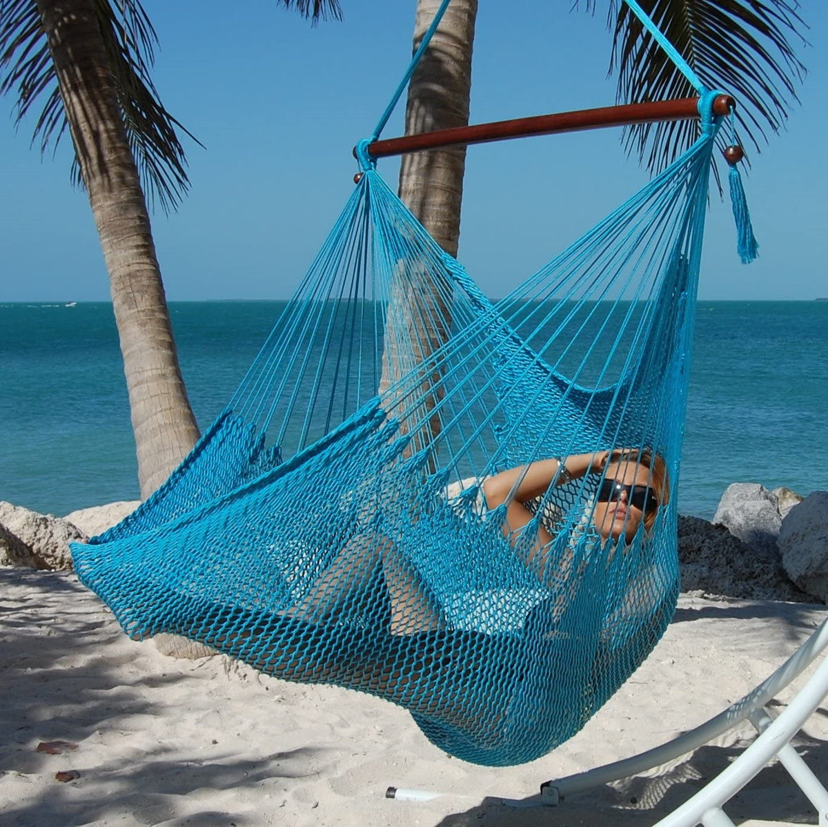 Woman reclining in a light blue Large Caribbean Hammock Chair on a beach with palms and turquoise water behind