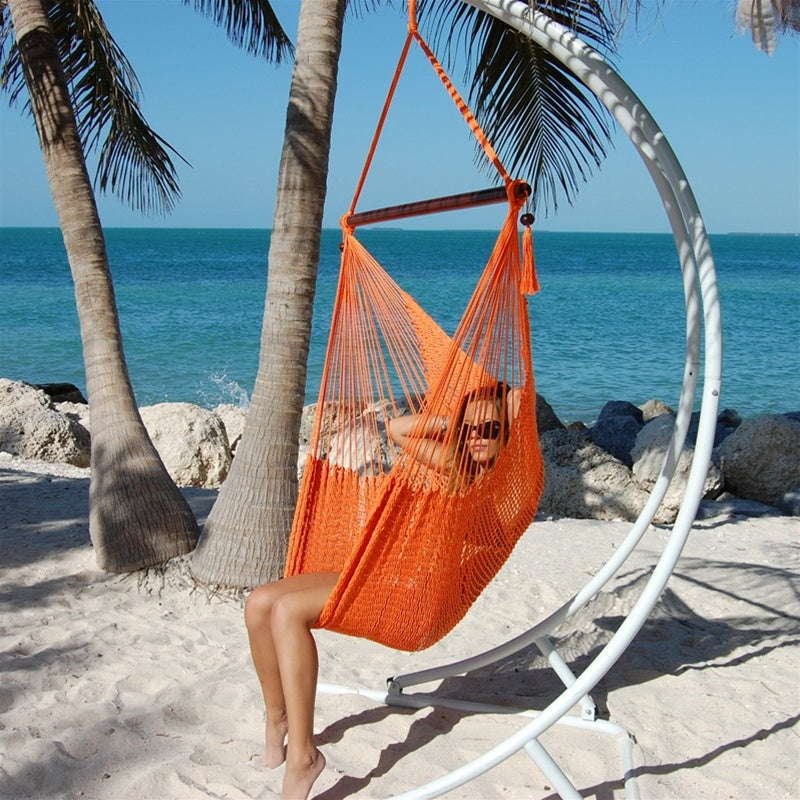 Woman relaxing in an orange Large Caribbean Hammock Chair on a white arc stand beneath palm trees by the ocean