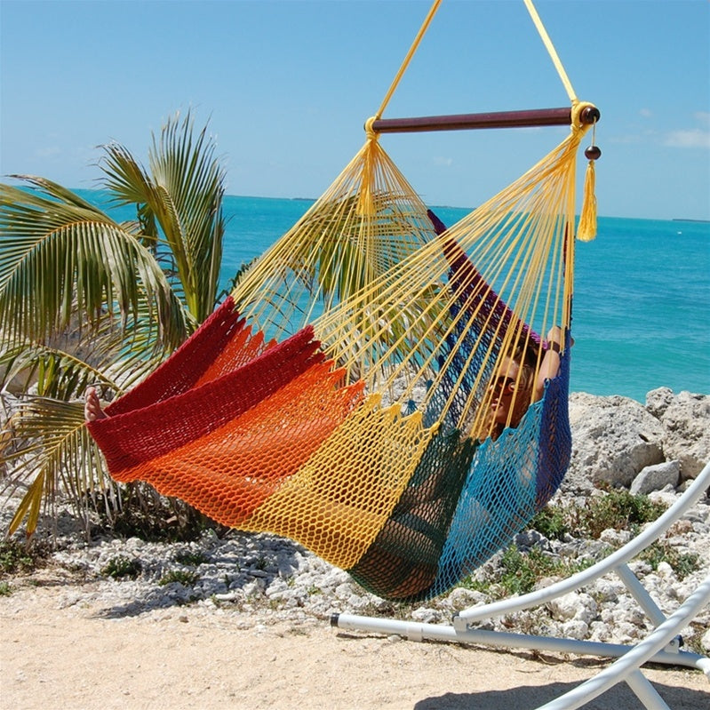 Woman reclining in a rainbow Large Caribbean Hammock Chair with yellow end cords, hung from a white arc stand on a sandy beach