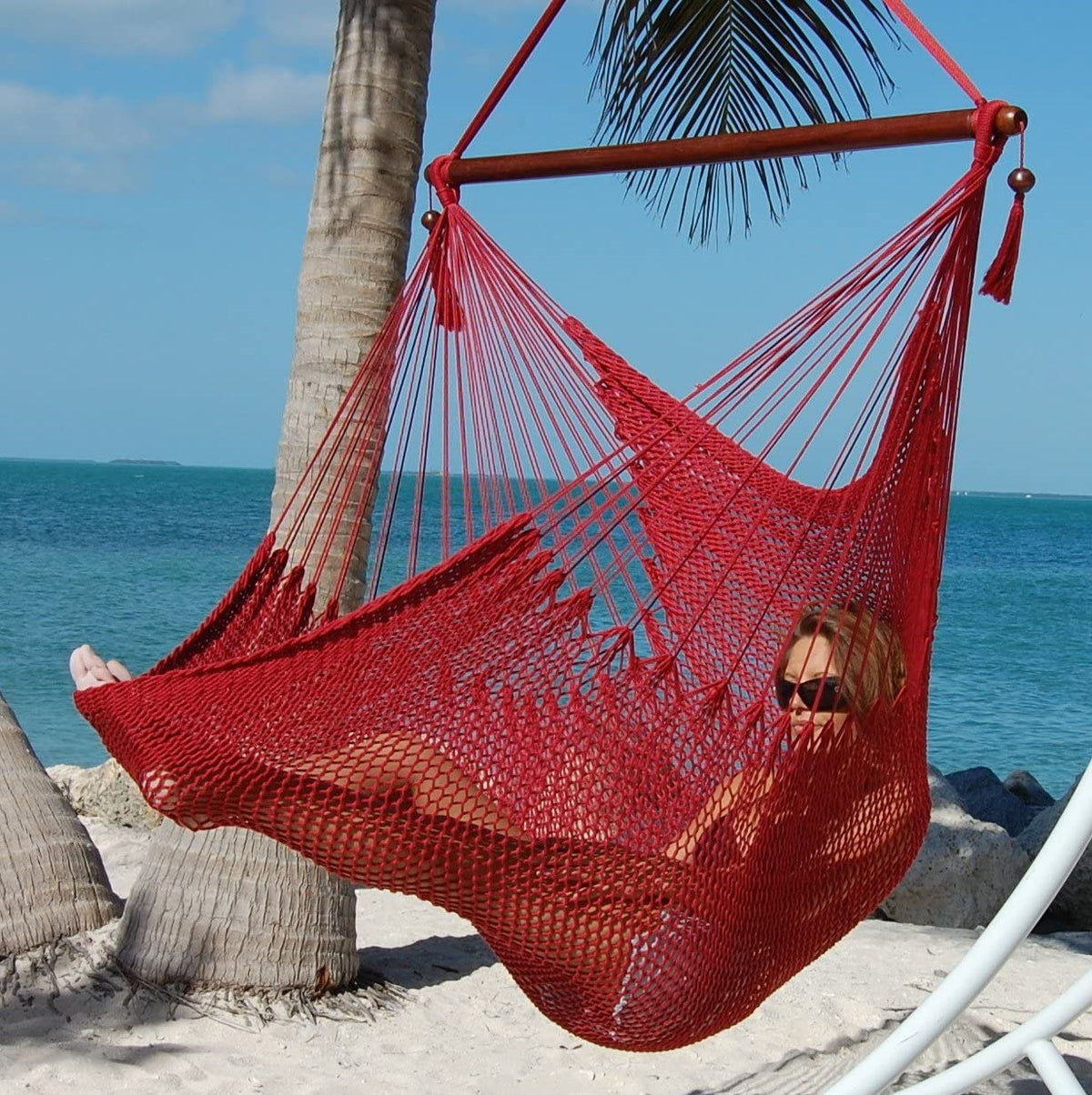 Woman reclining in a red Large Caribbean Hammock Chair on a white arc stand beside palm trees and the ocean