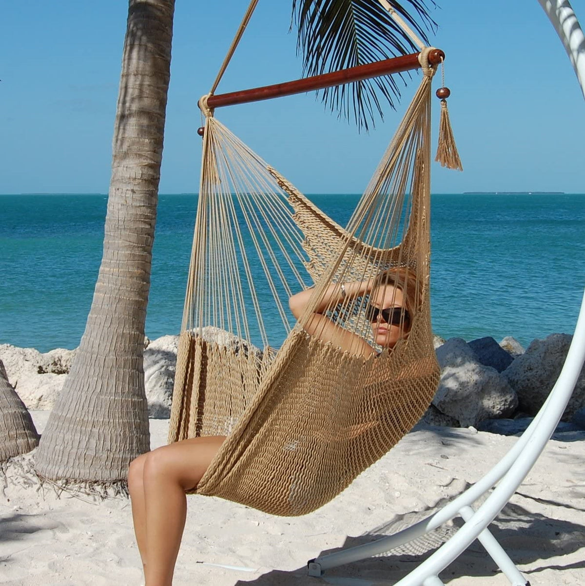 Woman relaxing in a tan Large Caribbean Hammock Chair on a white arc stand beneath palm trees by the ocean